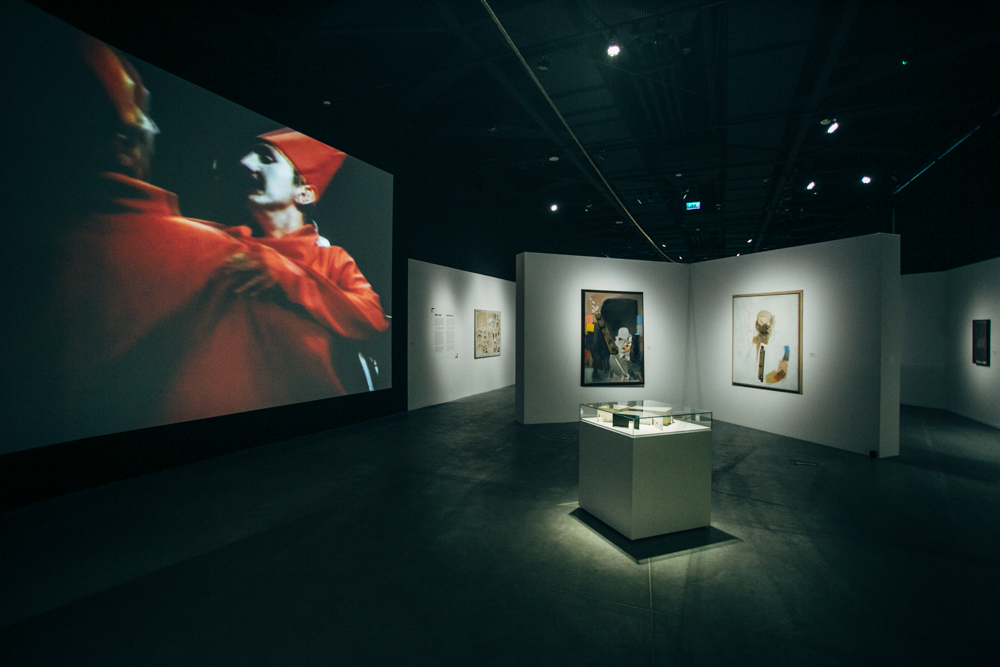 Wnętrze wystawy, na ściankach działowych wiszą obrazy, na ścianie wyświetla się obraz dwóch tańczących mężczyzn