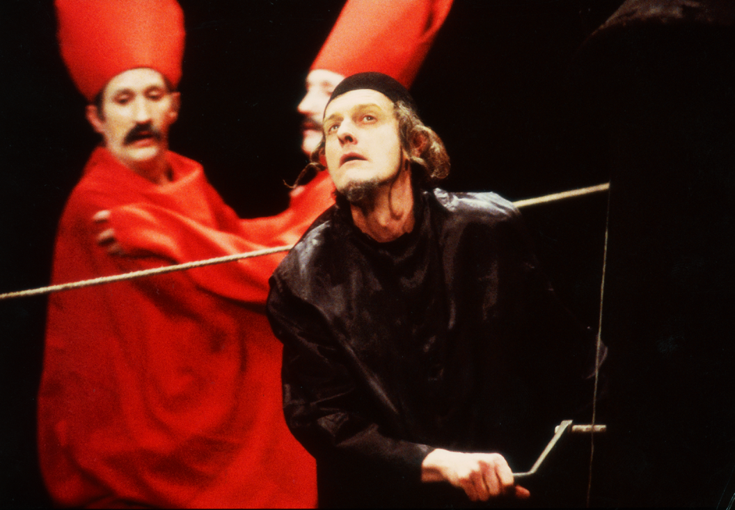 Zdjęcie ze spektatklu; mężczyzna w czarnych szatach, w tle para aktorów w czerwonych ubraniach