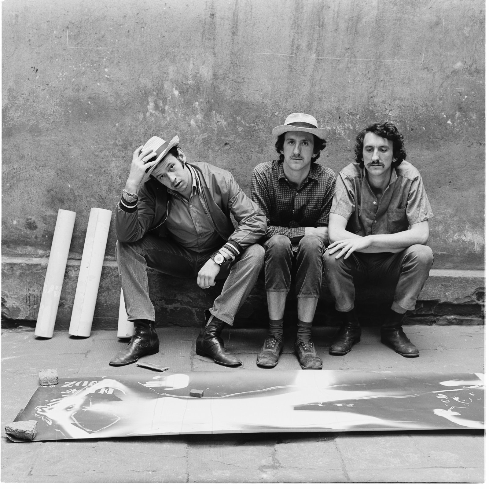 3 mężczyzn siedzących na murku; przed nimi na ziemi leży element wystawy
