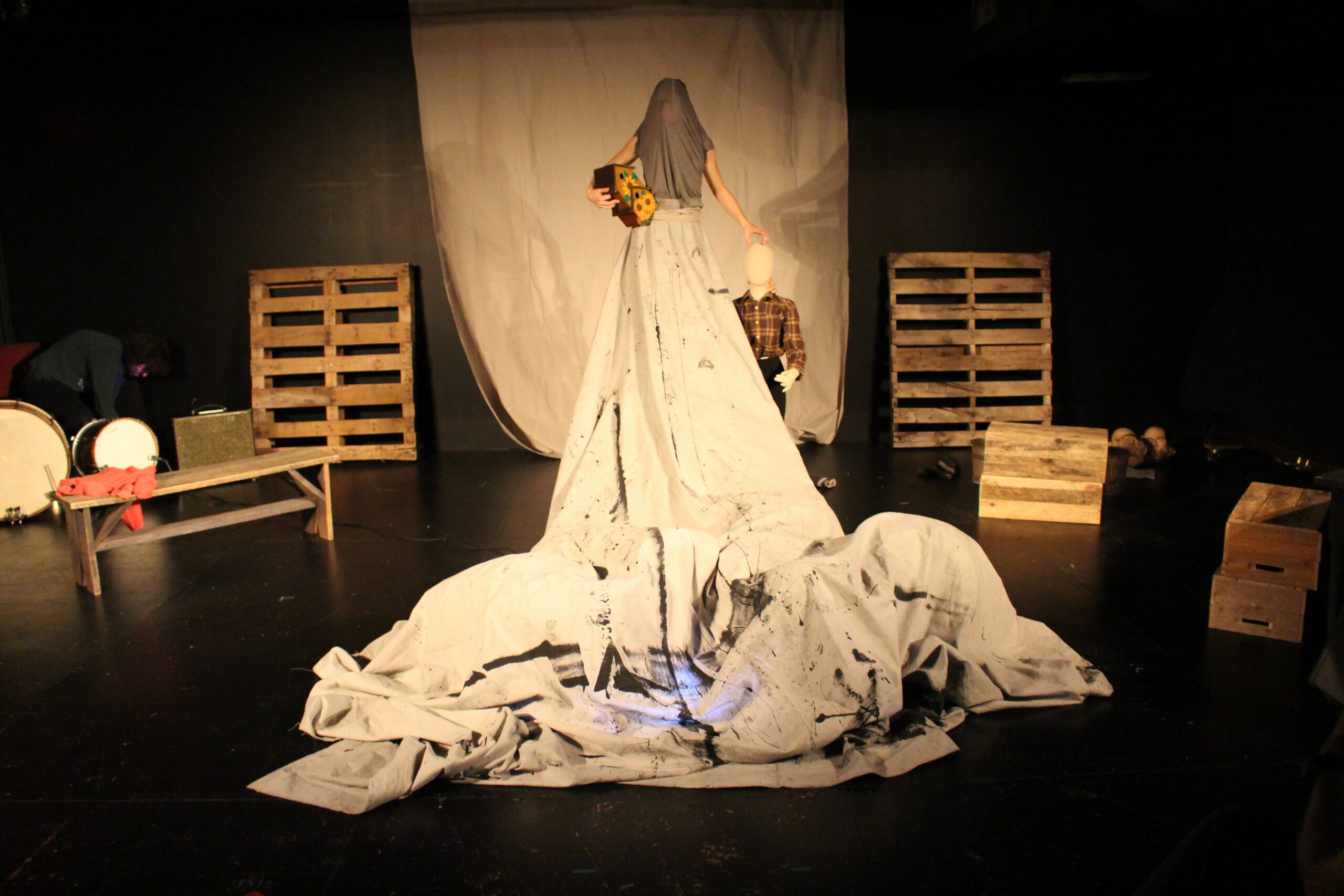 Zdjęcie scenografii; nienaturalnie wysoka sylwetka w sukni z płótna, drewniane skrzynki i palety