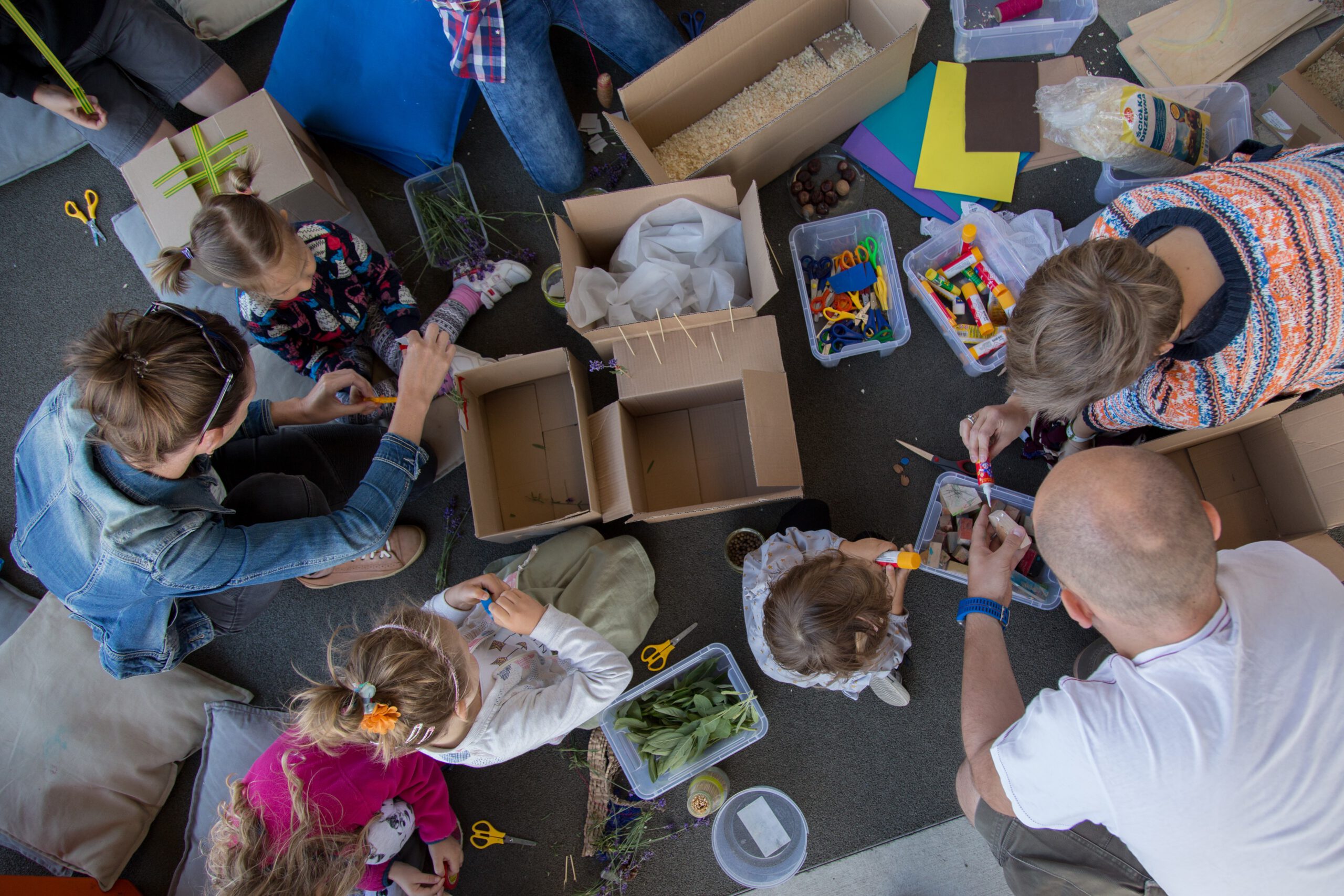 Na zdjęciu grupa dorosłych i dzieci pracująca z nożyczkami, klejem i kartonami.