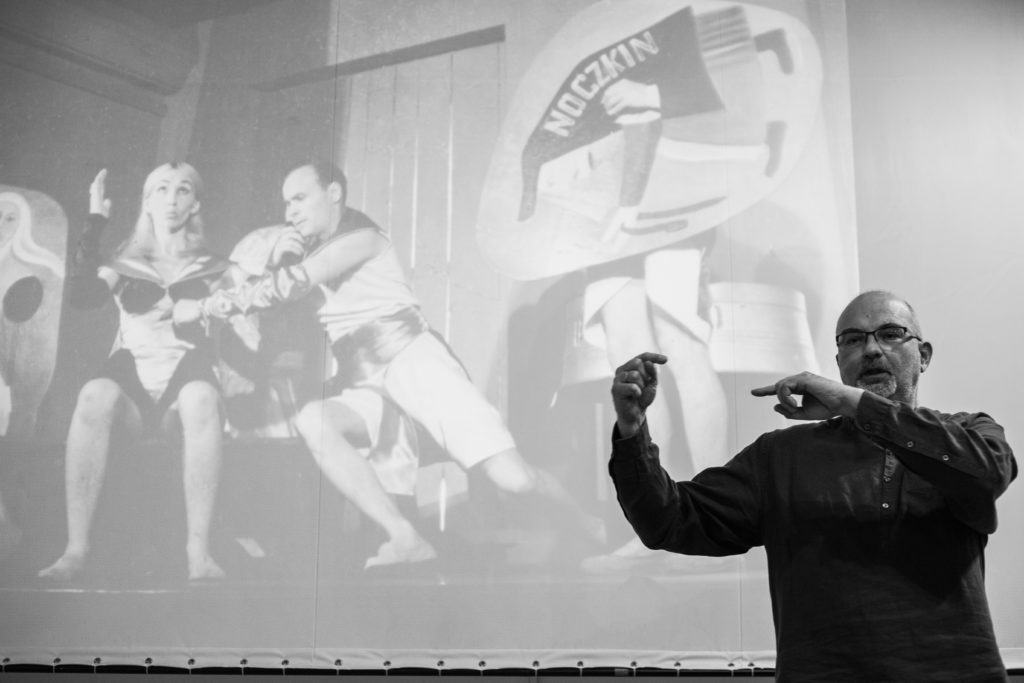 Na czarno białym zdjęciu Dariusz Kosiński wygłasza wykład, w tle zdjęcia ze spektaklu
