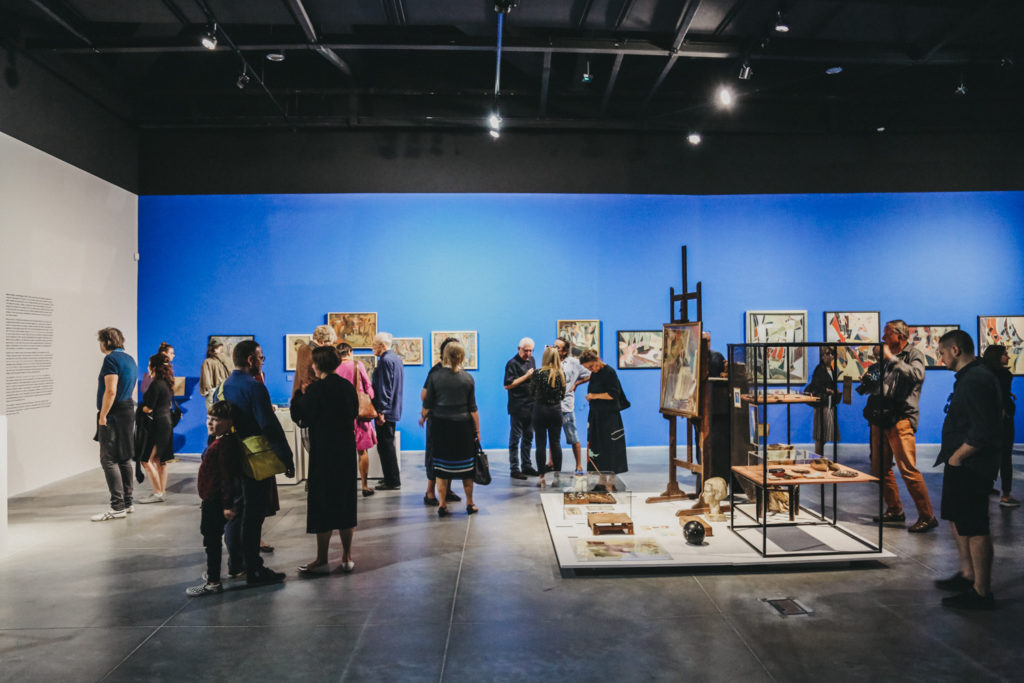Zdjęcie: widzowie zwiedzają wystawę Jaremianki. Kadr skierowany jest na bibeloty z pracowni artystki i przeglądające je osoby stojące na tle obrazów. 