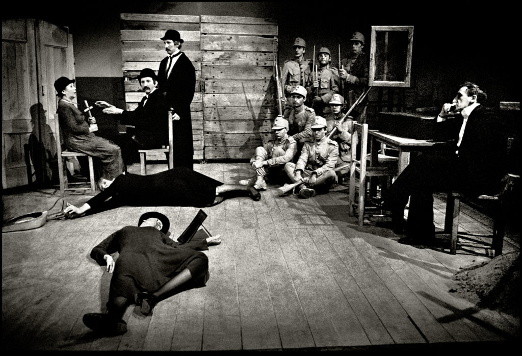 Czarno-biała fotografia przedstawia scenę ze spektaklu. Z prawej strony Tadeusz Kantor siedzi przy stole. W głębi grupa żołnierzy ustawiona jak do pamiątkowego zdjęcia. Z lewej strony aktorzy siedzą na krzesłach. Przed nimi na drewnianej podłodze leżą dwie osoby w dziwnych pozach. 