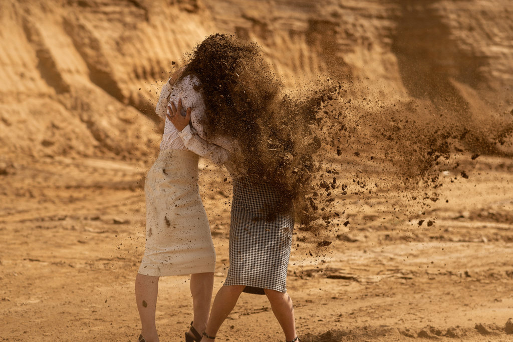 Na zdjęciu widać dwie przytulone kobiety zasłaniające się przed piaskiem. 