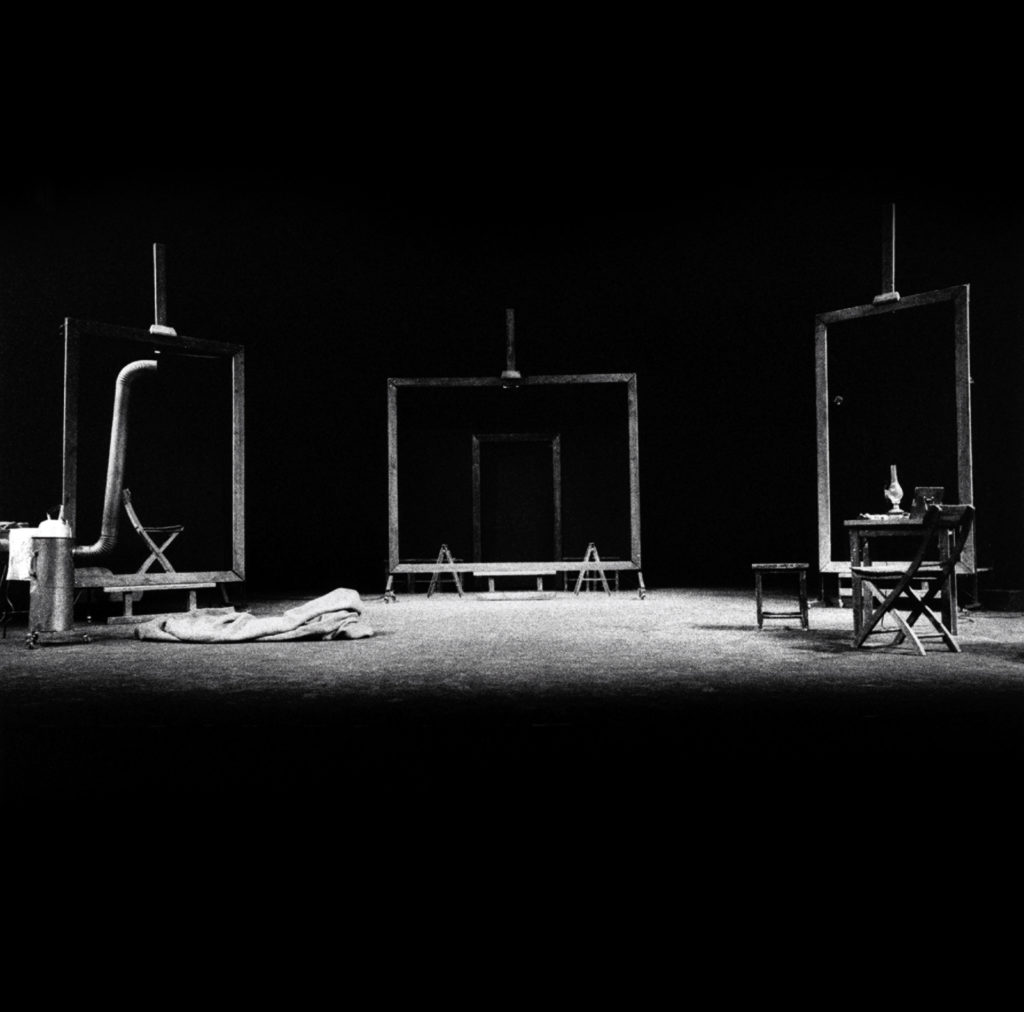 Czarno-białe zdjęcie przedstawia scenografię spektaklu "Dziś są moje urodziny": trzy puste ramy obrazów wysokości dorosłego człowieka ustawione w półkolu, obok nich różne meble: stolik, krzesła, piecyk-koza.