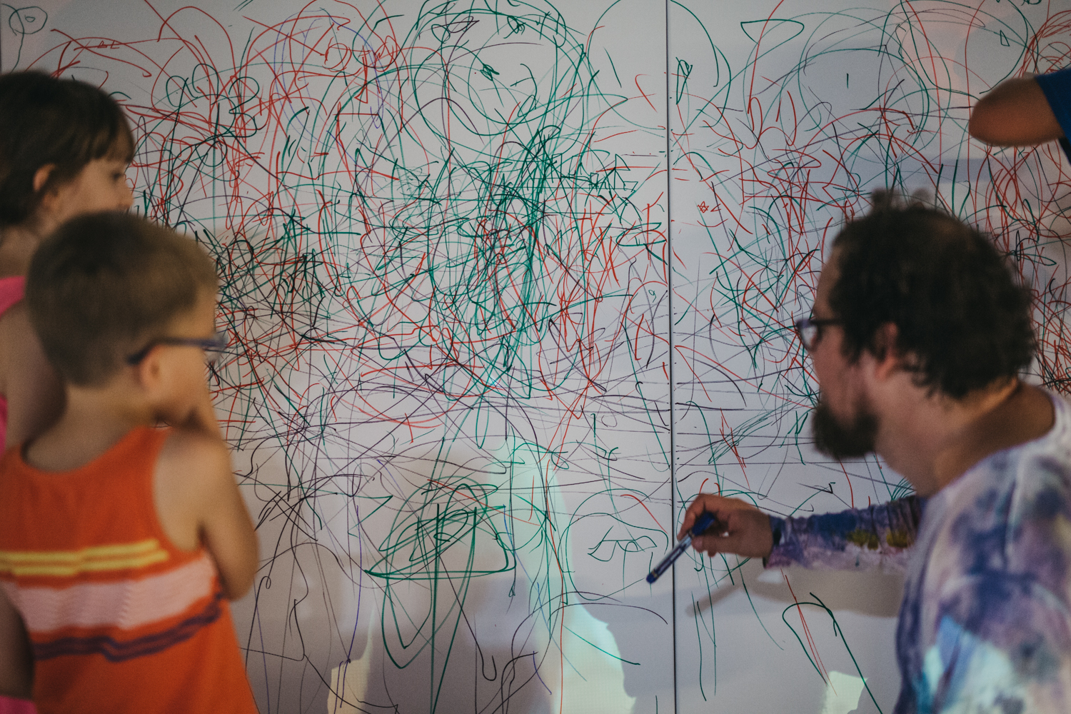 Na zdjęciu prowadzący wraz z dziećmi rysuje po podobraziu. Płótno pokryte jest gąszczem zielonych i czerwonych linii.