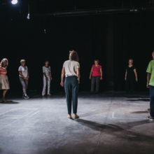 Stojąca w kręgu w sali teatralnej grupa kilkunastu osób w różnym wieku