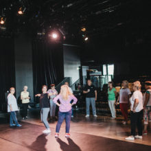 Grupa osób stoi w półokręgu w sali teatralnej. Dwie osoby tańczą pośrodku.