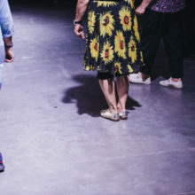 Osoba w sukience w kwiaty i w srebrnych butach stoi w sali teatralnej