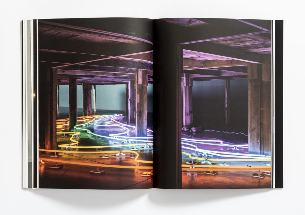 [Kolorowe zdjęcie książki "Zapraszanie. Sarkis-Kantor". Zdjęcie przedstawia drewniany stół, pod nim neon. Neon składa się z kolorowych linii.]