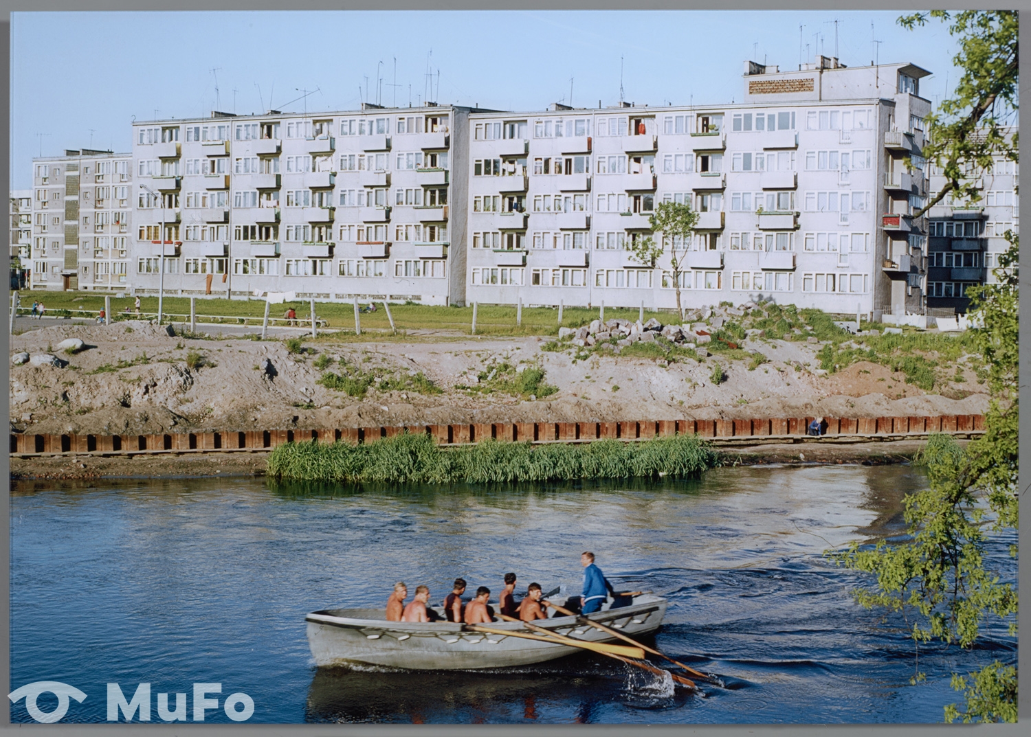 Kolorowe zdjęcie, które przedstawia bloki i rzekę. Płyną po niej w łódce mężczyźni.