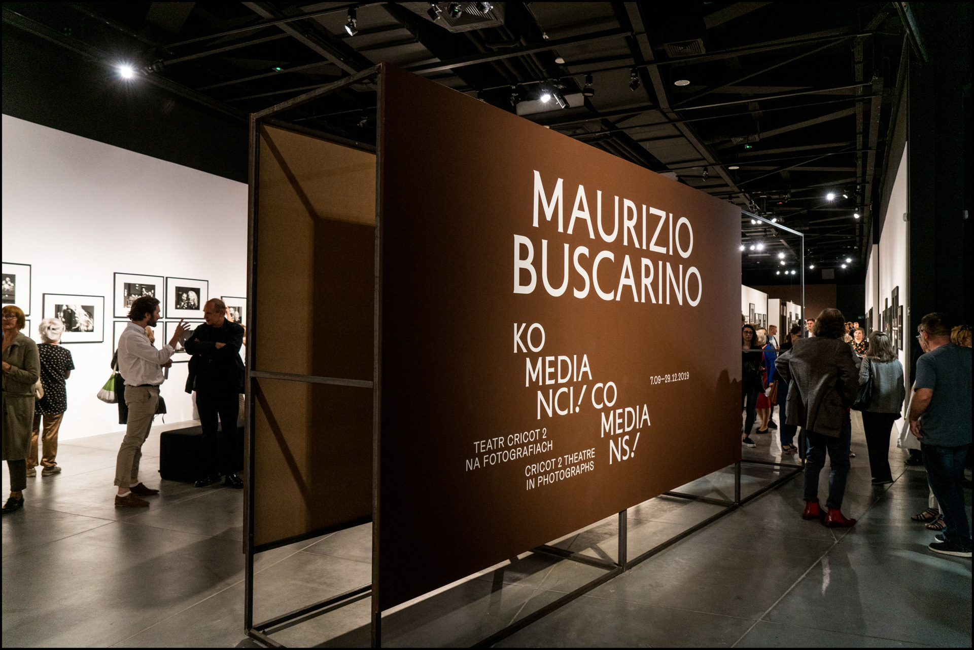 Zdjęcie przedstawia ujęcie z wernisażu wystawy fotografii "Maurizio Buscarino. Komedianci!". Na zdjęciu widać kobiety i mężczyzn oglądających wystawę. Na środku sali ustawiony jest brązowy baner z białymi napisami "Maurizio Buscarino. Komedianci!".