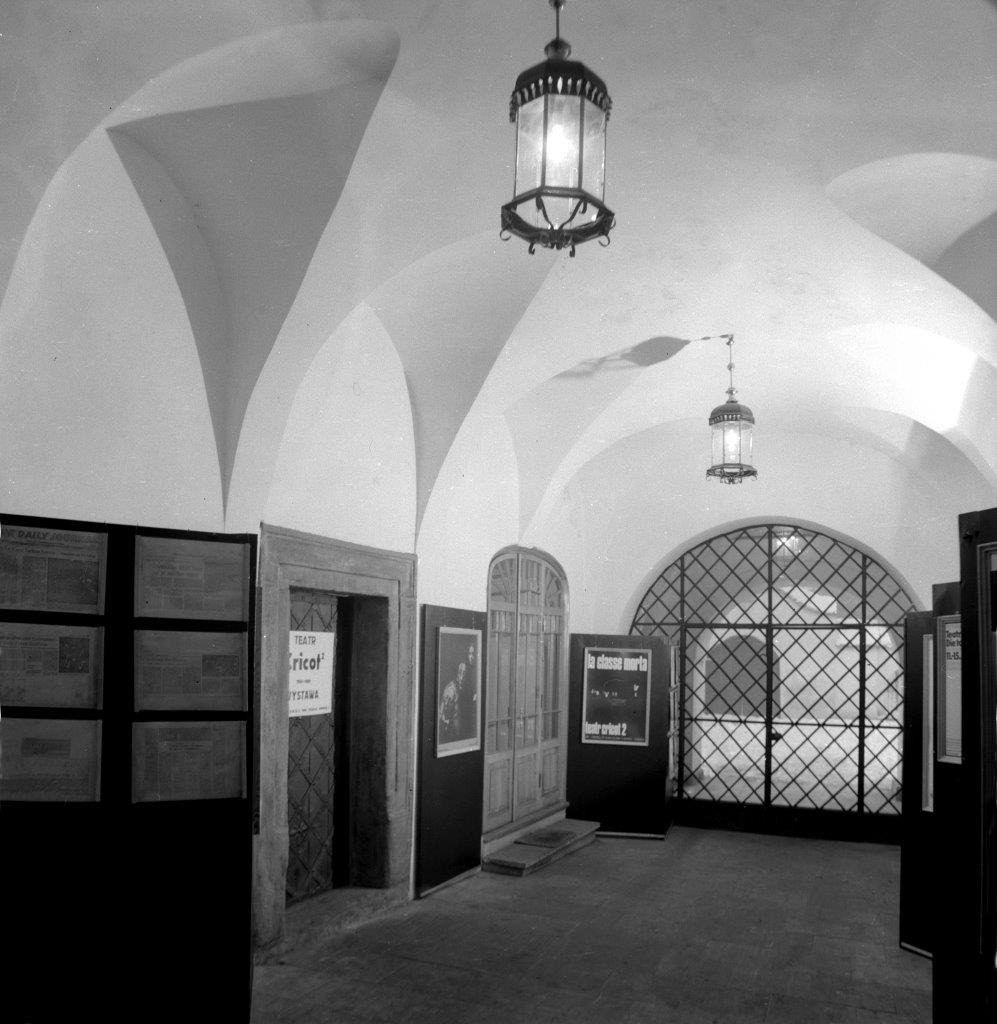 [Czarno-białe zdjęcie przedstawiające wnętrze Ośrodka Teatru Cricot 2 w 1980 roku. Widać na nim korytarz, drewniane drzwi, plakaty Teatru Cricot 2. Na końcu korytarza znajduje się żelazna brama.]
