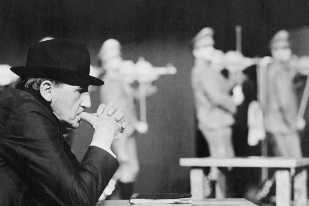 Czarno białe zdjęcie ze spektaklu Nigdy tu już nie powrócę, na pierwszym planie Tadeusz Kantor siedzi przy stole, na głowie ma czarny kapelusz