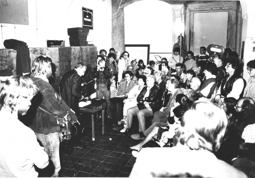 [czarno-białe zdjęcie, przedstawiające Kantora oraz tłum dziennikarzy w pomieszczeniu na Kanoniczej.]