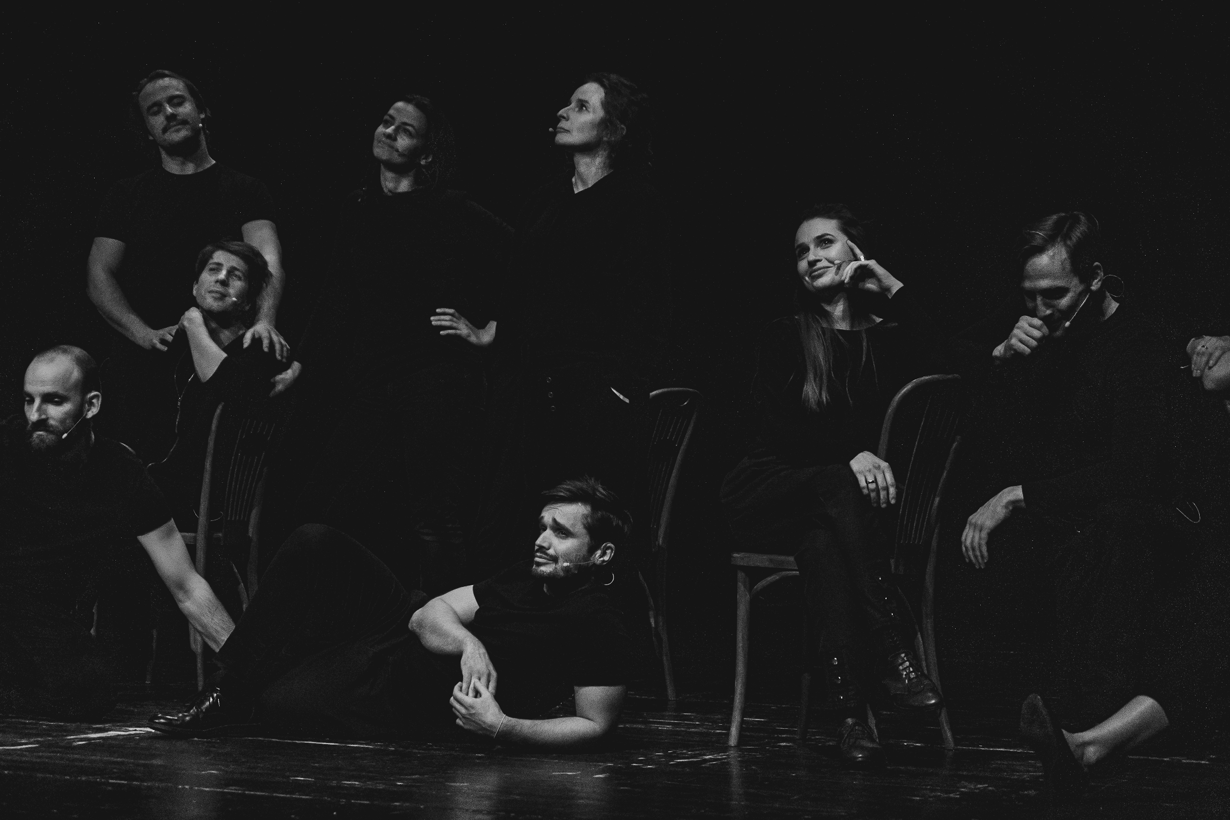 Czarno białe zdjęcie. Grupa dorosłych osób w czarnych ubraniach stoi i leży na ciemnej scenie