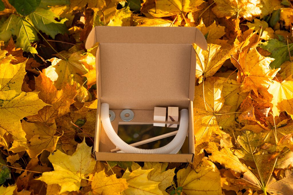 Kartonowe pudełko z drewnianymi klockami, rurką i metalowymi kółkami leży pośród jesiennych liści.