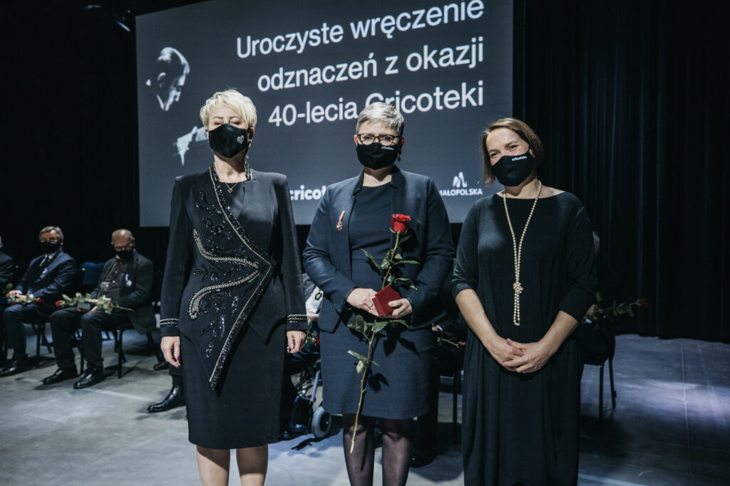 Zdjęcie przedstawia trzy osoby. W środku stoi Agnieszka Oprządek. Trzyma czerwoną różę. Do czarnego żakietu ma przypiętą odznakę “Zasłużony dla Kultury Polskiej”. Po lewej stoi Iwona Gibas. Po prawej stoi Natalia Zarzecka.