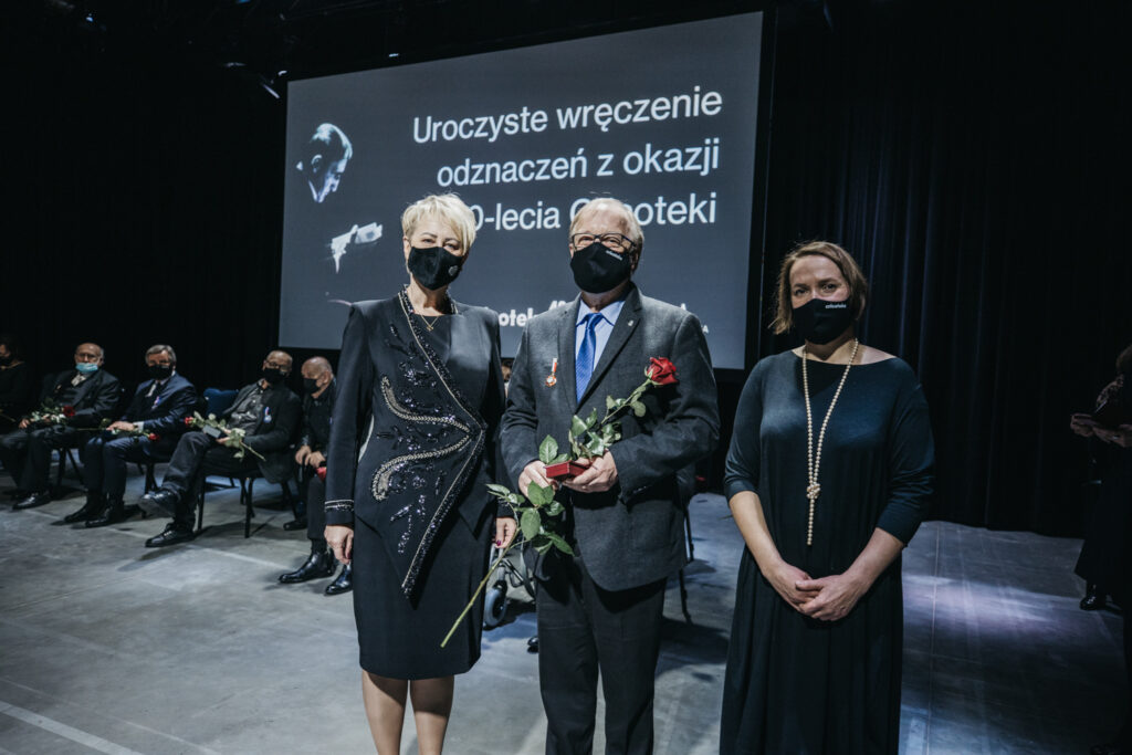Na zdjęciu są trzy osoby. W środku stoi Krzysztof Pleśniarowicz. Trzyma czerwoną różę. W klapę marynarki ma wpiętą odznakę “Zasłużony dla Kultury Polskiej”. Po lewej stronie stoi Iwona Gibas. Po prawej stoi Natalia Zarzecka. W tle znajdują się inni nagrodzeni, którzy siedzą na krzesłach.