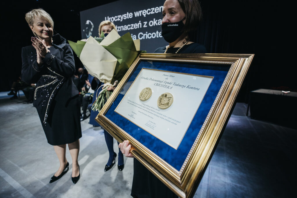 Natalia Zarzecka trzyma ramę z Medalem Polonia Minor dla Cricoteki. Jest uśmiechnięta i wyraźnie wzruszona. W ręce trzyma bukiet kwiatów. Po lewej stoi Iwona Gibas. Klaszcze w dłonie.