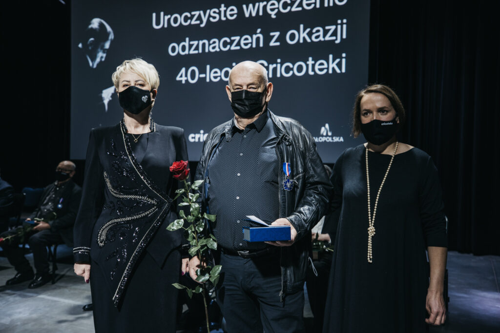 Na zdjęciu są trzy osoby. W środku Jacek Maria Stokłosa. Ma przypięty Srebrny Medal Gloria Artis. Trzyma czerwoną różę. Po lewej stoi Iwona Gibas. Po prawej stoi Natalia Zarzecka.