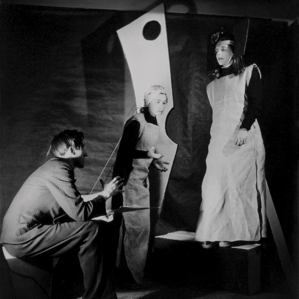 Archiwalne zdjęcie. Na pierwszym planie siedzi Tadeusz Kantor, spoglądający w stronę dwóch kobiet w sukniach. Za nimi góruje wysoki, geometryczny kształt z okrągłą dziurą - okiem.