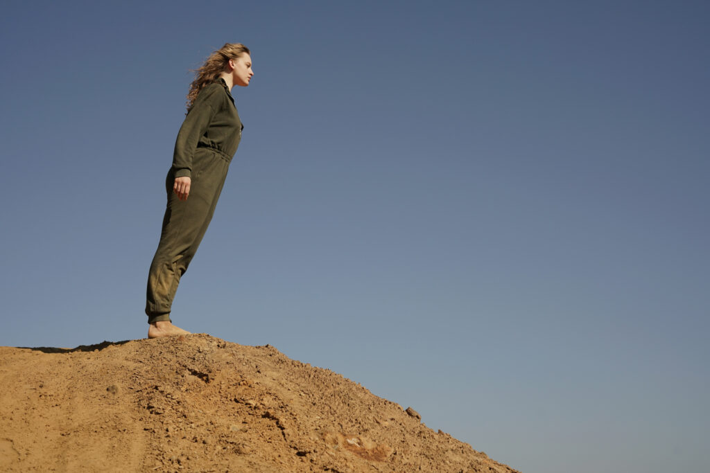 Kobieta w kombinezonie roboczym stoi na nasypie z piasku. Jest mocno wychylona do przodu, jakby za moment miała upaść