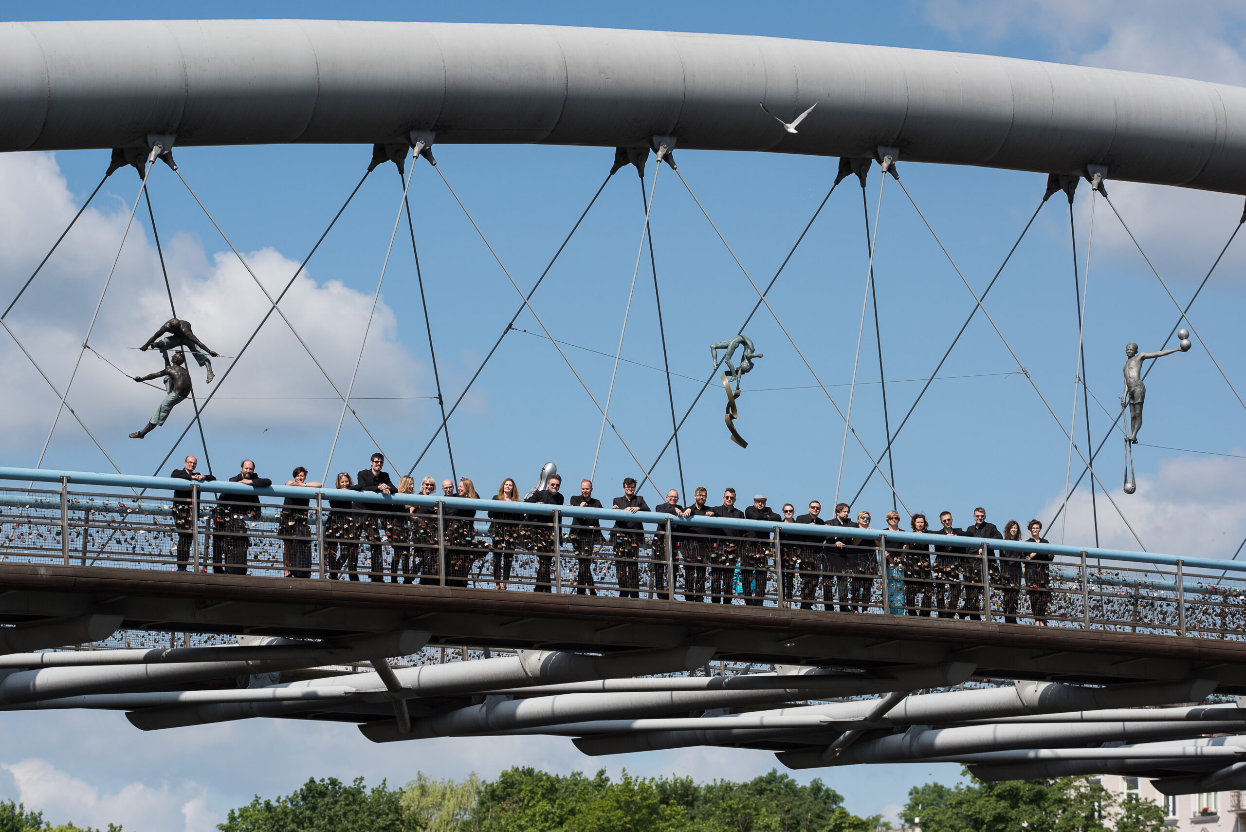 Grupa osób ubranych na czarno stojąca na moście. Nad nimi powieszone instalacje na moście.