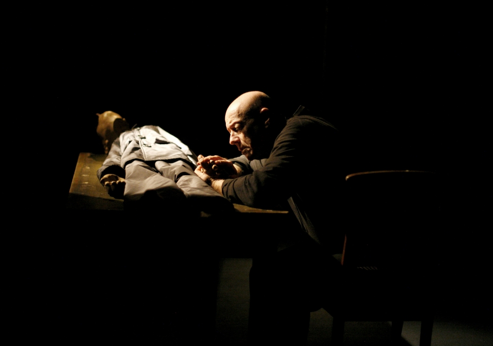 Łysy mężczyzna siedzi w ciemnym pomieszczeniu. Pochyla się nad lalką, która leży przed nim na stole