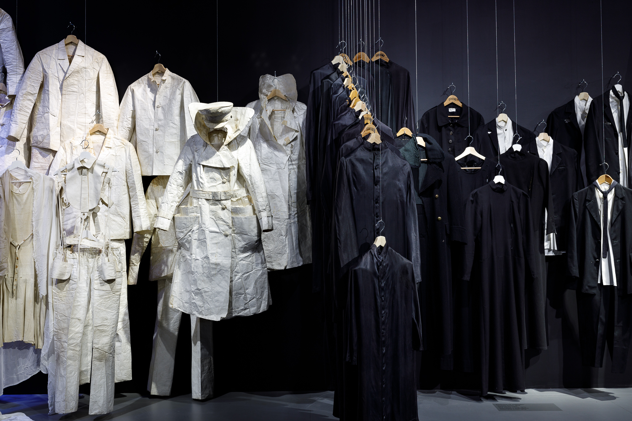 Zdjęcie z wystawy "Osobiste. Kostiumy teatralne Kantora". Z lewej strony wisi kilka białych kostiumów. Po prawej wiszą czarne szaty na drewnianych wieszakach