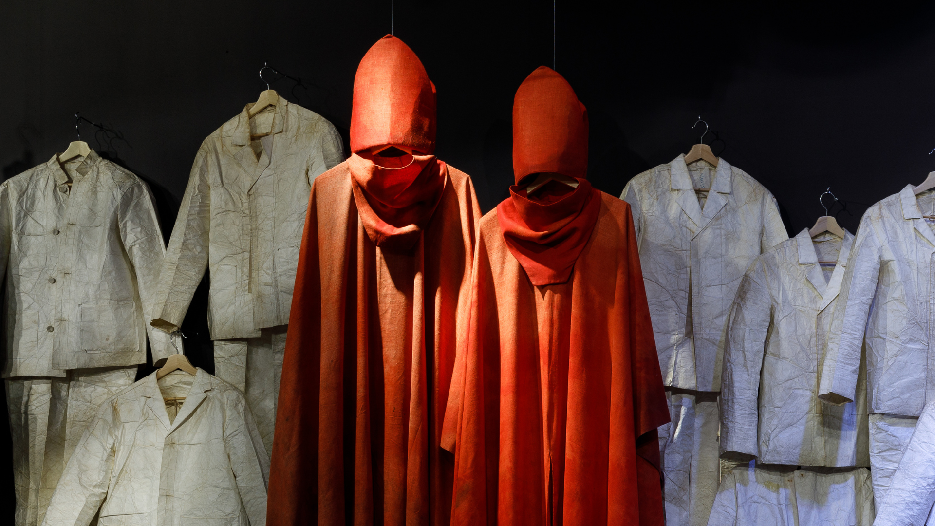 Zdjęcie kostiumów wiszących koło siebie. W centrum znajdują się dwa czerwone kostiumy biskupów. Wokół nich wiszą białe, przypominające mundury ubrania.