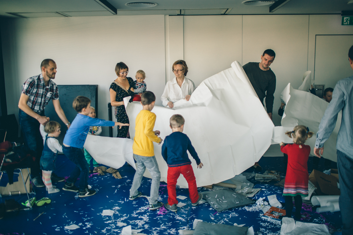 Zdjęcie przedstawia grupę osób dorosłych i dzieci bawiących się dużym białym kartonem, wśród ścinków papieru