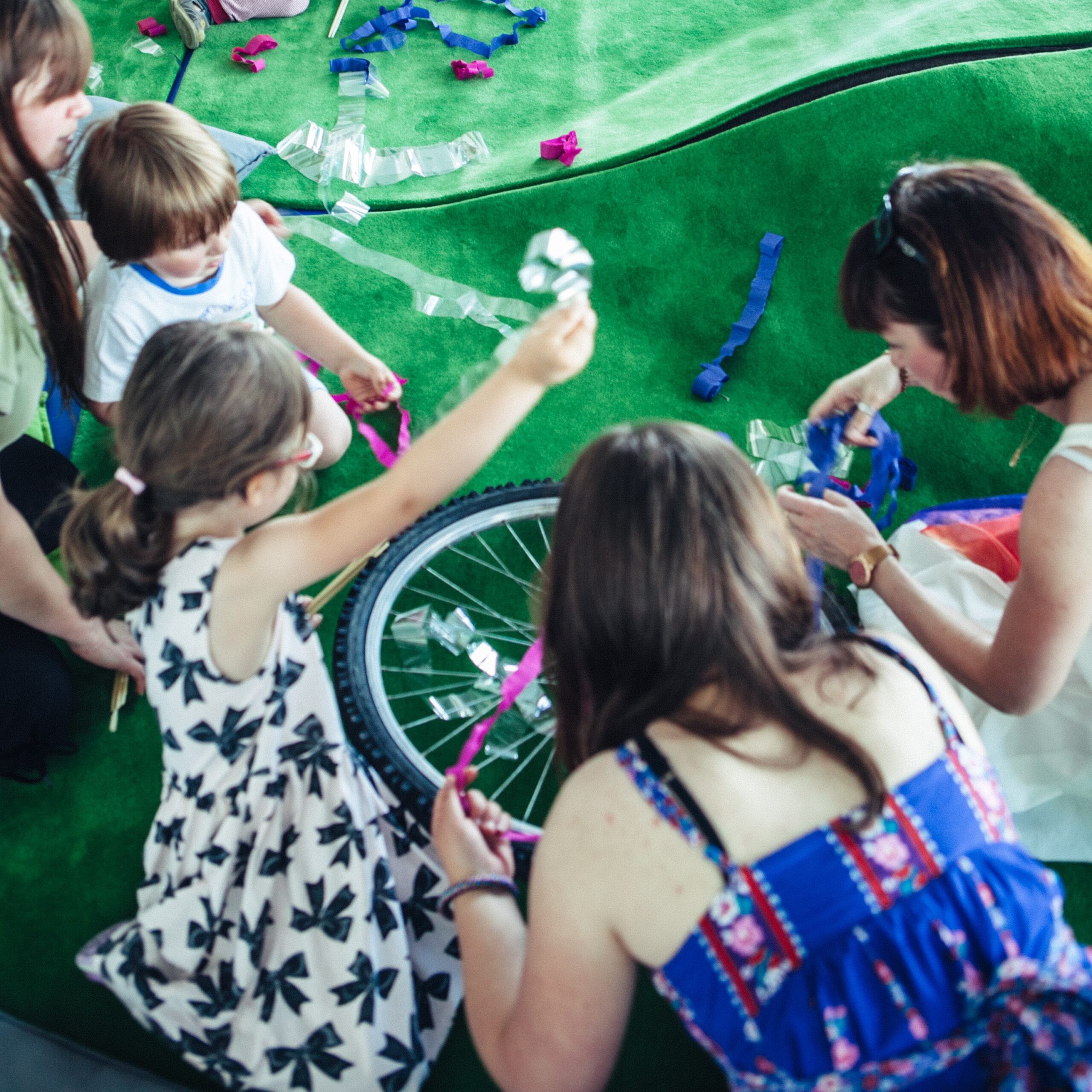 Fotografia przedstawia dzieci siedzące za zielonek wykładzinie i bawiące się kolorowymi bibułami