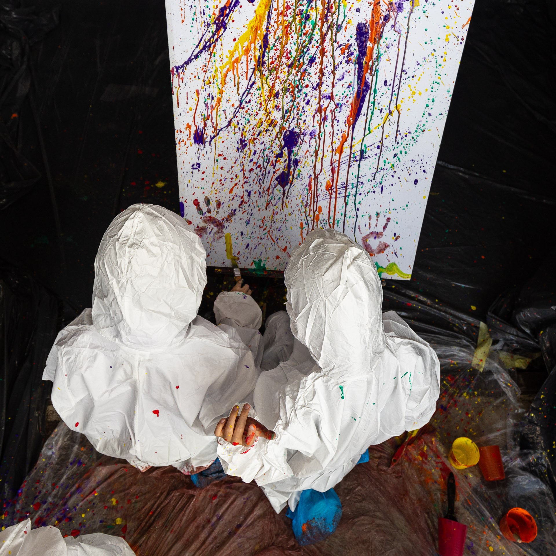 Zdjęcie przedstawia dwoje dzieci w białych kombinezonach chlapiących farbą na biały karton