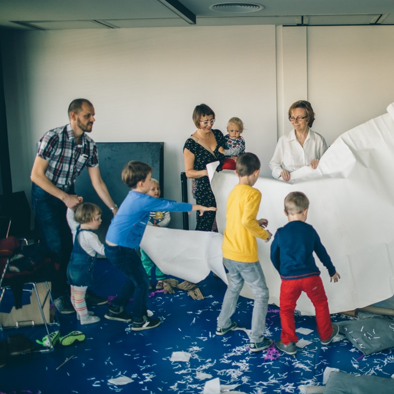 Na fotografii widzimy osoby dorosłe i dzieci bawiące się wielkim arkuszem papieru w kolorze białym
