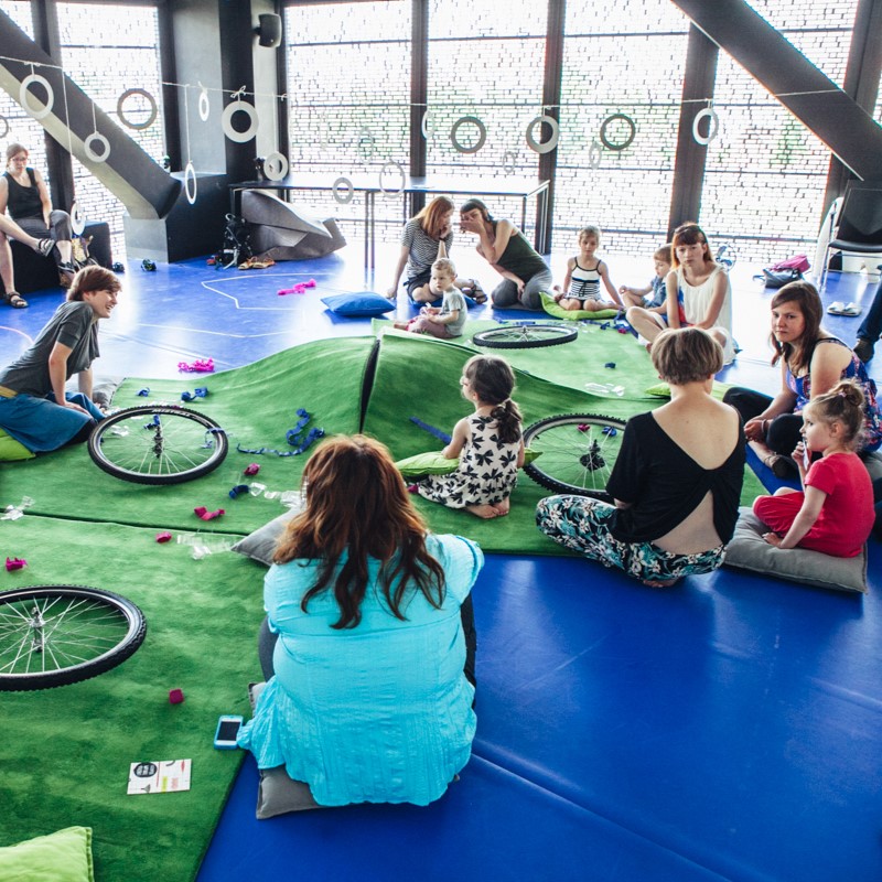 Dzieci i dorośli siedzą na niebieskiej podłodze, wokół zielonej wykładziny, na której leżą różne części - np. koła rowerowe.