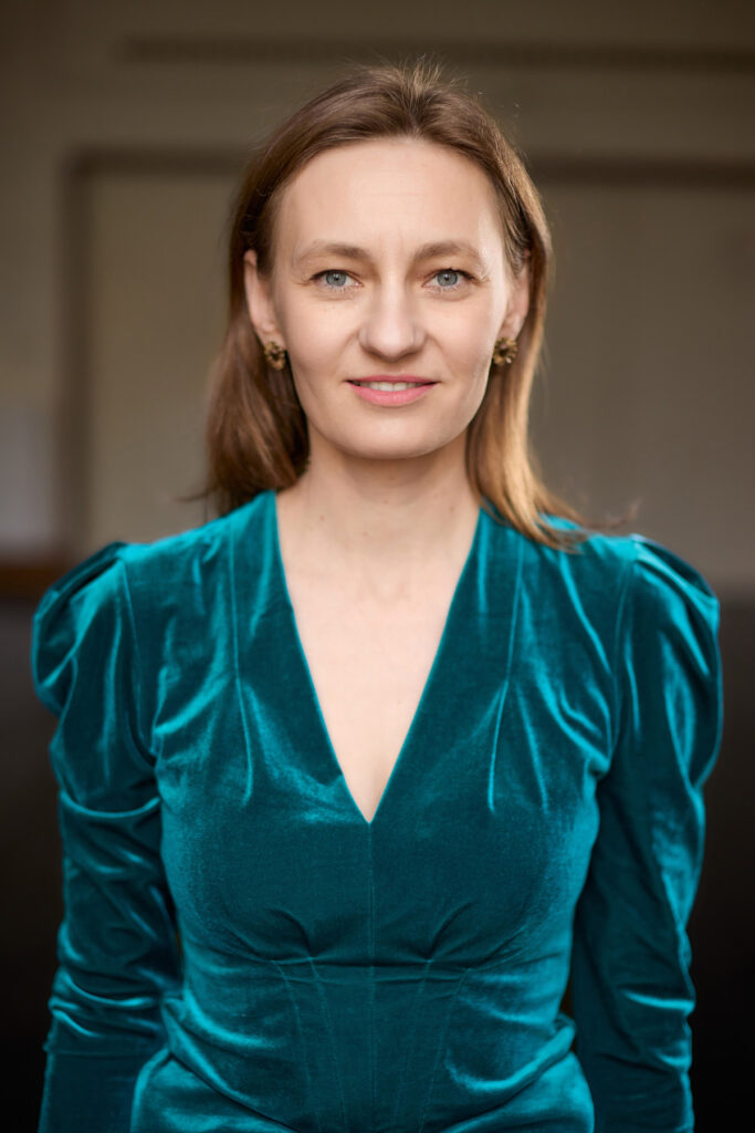 Na ciemnym tle stoi młoda uśmiechnięta kobieta z długimi włosami koloru ciemny blond, ubrana w sukienkę koloru cyjanowego.