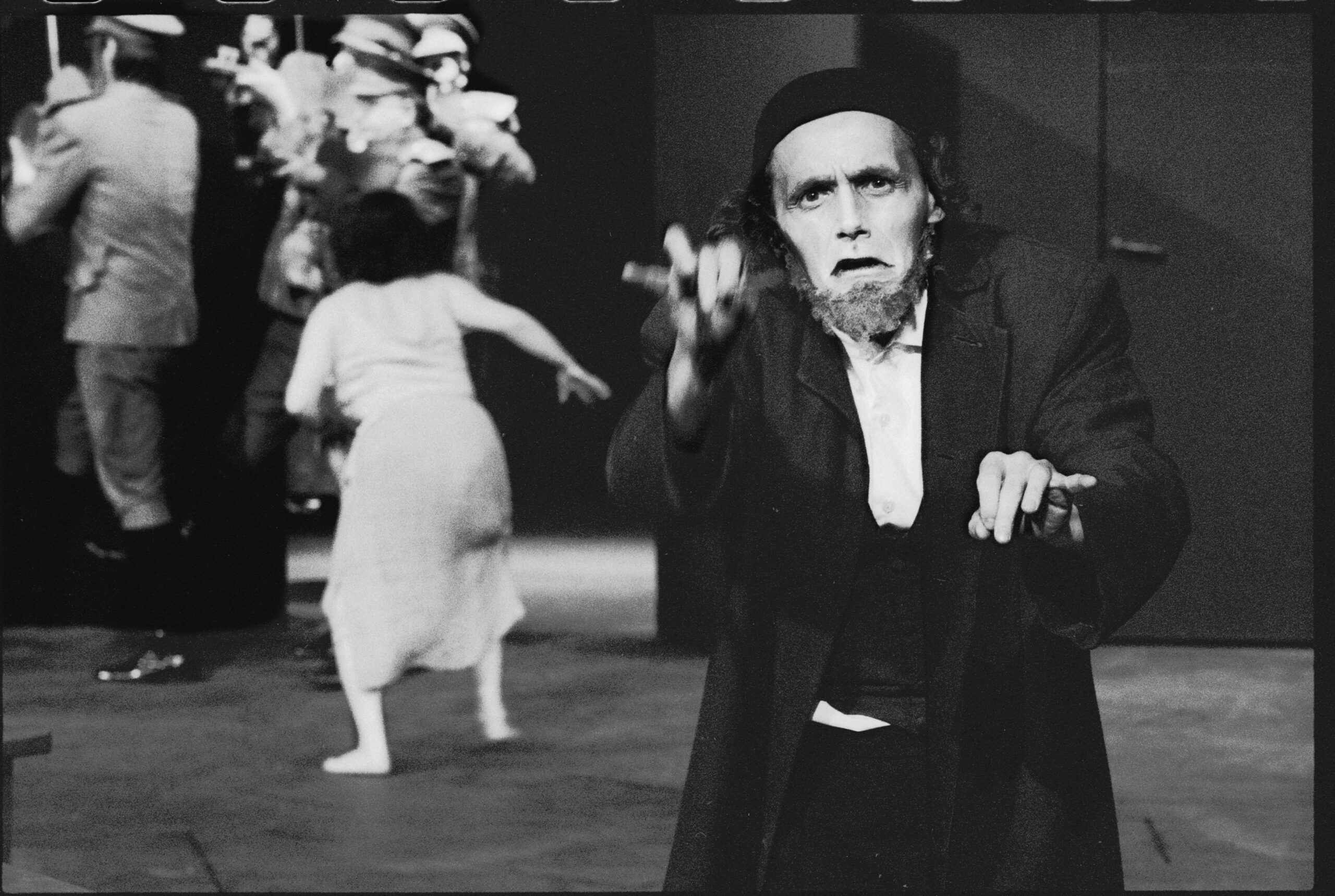 Czarno-biała fotografia pokazuje scenę teatralną z mężczyzną stojącym na pierwszym planie. Ma na sobie ciemny płaszcz i czapkę, a jego twarz wyraża silne emocje, gdy unosi rękę w geście. W tle widać rozmyte postacie w ruchu, co sugeruje trwający spektakl.