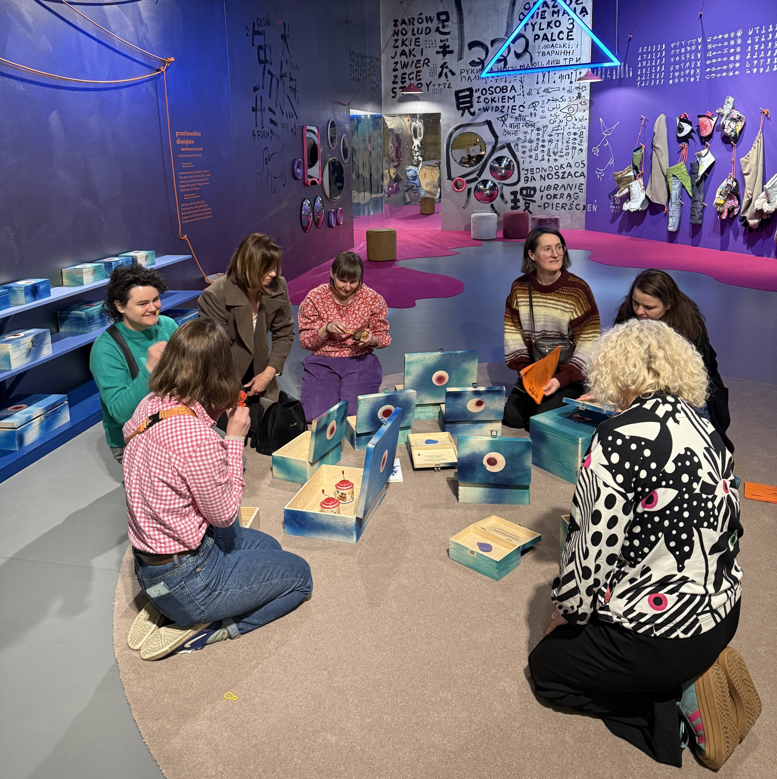 Grupa siedmiu osób siedzi w kręgu na podłodze w kolorowej, artystycznej przestrzeni wystawowej. Uczestnicy skupiają się na interaktywnych obiektach — drewnianych pudełkach i elementach z okrągłymi wzorami — które leżą na środku. W tle widoczne są ściany w odcieniach fioletu z rysunkami, napisami i instalacjami artystycznymi, a także zawieszone elementy dekoracyjne i lustra. Atmosfera wygląda na kreatywną i warsztatową.