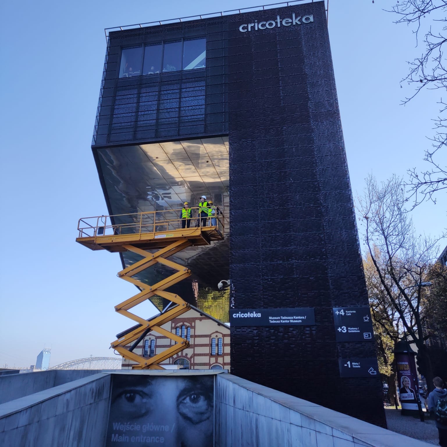 Na zdjęciu widoczny jest budynek Cricoteki sfotografowany od frontu, z charakterystyczną ciemną elewacją i dużą, wysuniętą nad wejściem bryłą. Na podnośniku nożycowym, uniesionym do wysokości zadaszenia, stoją trzy osoby w kaskach i kamizelkach odblaskowych, prowadzące prace przy spodniej części konstrukcji. W tle widać bezchmurne niebo, drzewa oraz fragment zabytkowego budynku i mostu, a na pierwszym planie znajduje się wejście do muzeum z dużym czarno-białym portretem Tadeusza Kantora.
